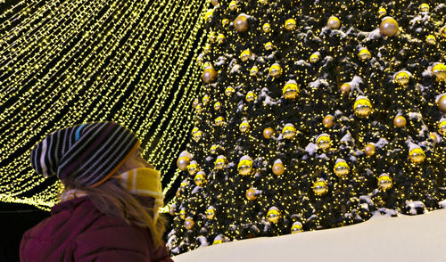 Young woman in knitted yellow scarf looking at christmas tree decorated golden balls, garlands light