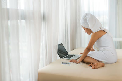 Woman wrapped in towel while using laptop on bed at home