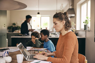 Side view of woman using laptop while family in background at home
