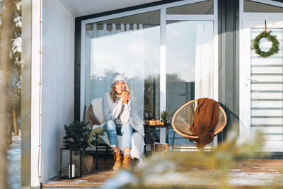 Smiling woman in winter clothes with coffe cup sitting near country house in winter