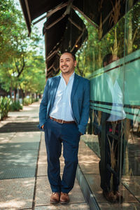 Full body happy hispanic mature male manager in suit holding hands in pockets and looking away with smile while leaning on glass wall on sunny day on city street