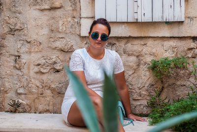 Portrait of young woman wearing sunglasses and white dress sitting against wall
