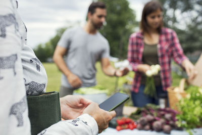 Woman using mobile phone while couple selling vegetables in background at urban garden