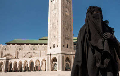 Woman in burka standing by mosque against clear blue sky