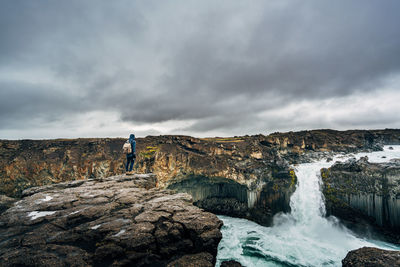 Hiker standing on cliff by waterfall against cloudy sky