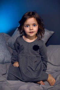 Girl in a gray dress sits on a bed with pillows in a bedroom with a blue wall