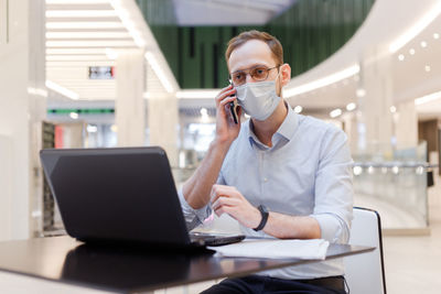 Businessman wearing mask talking on phone while sitting at mall