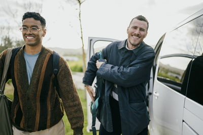 Smiling male friends with backpacks walking by van