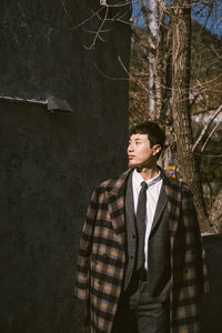 Young man looking away while standing against trees