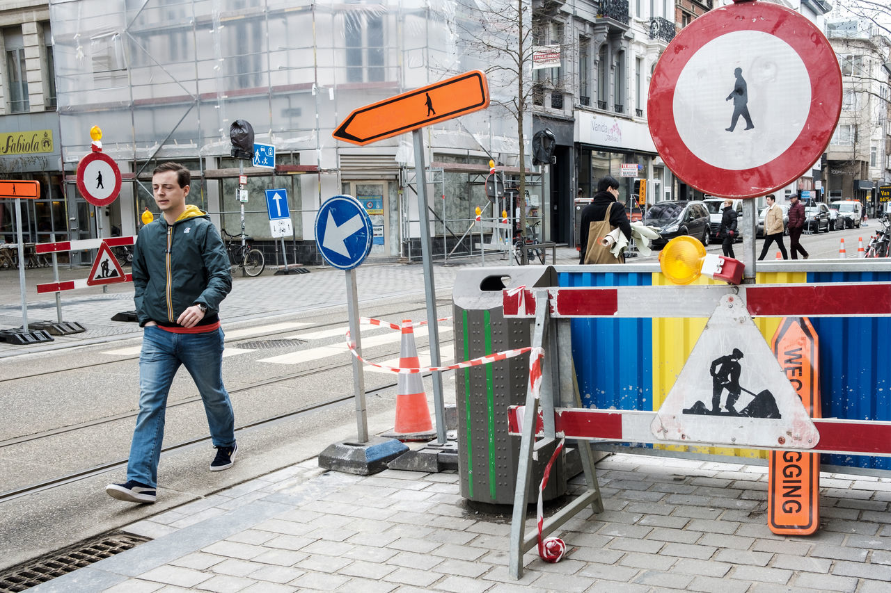 city, architecture, road sign, road, sign, building exterior, men, street, real people, day, transportation, built structure, casual clothing, communication, city life, mode of transportation, human representation, people, young adult, young men, guidance, outdoors