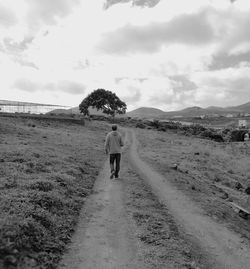 Rear view of man walking on dirt road against cloudy sky