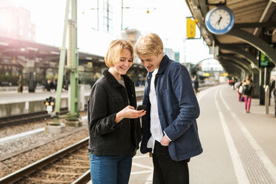 Smiling teenage girl showing mobile phone to young man while standing on railroad station platform