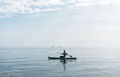 Young man from behind sitting on stand up paddle board on sea on sunny summer day, active lifestyle