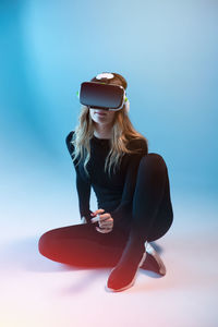Young unrecognizable blonde woman sitting wearing virtual reality headset on colorful blue neon illumination studio background