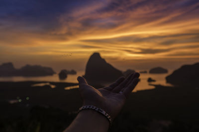 Cropped hand gesturing at beach against cloudy sky during sunset