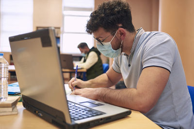 A boy in a mask is studying with a laptop computer in the library