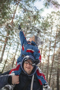 Happy little boy sits on the shoulders of his father and wants to reach the branch