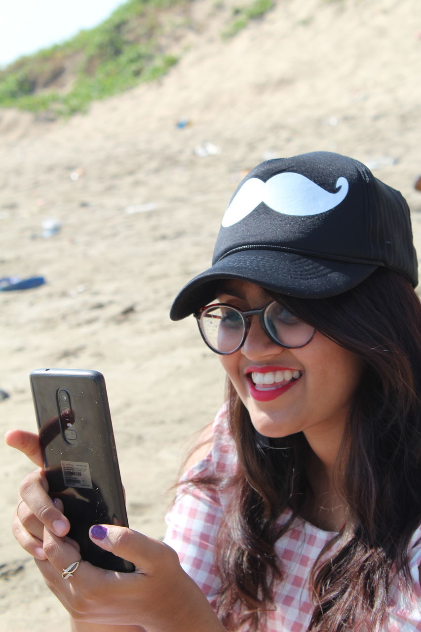 one person, mobile phone, real people, lifestyles, leisure activity, holding, wireless technology, technology, young adult, smart phone, young women, smiling, portable information device, portrait, hat, glasses, communication, focus on foreground, day, fashion, outdoors, hairstyle