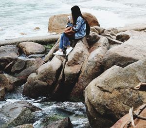 Woman holding disposable cup sitting on rocks by sea