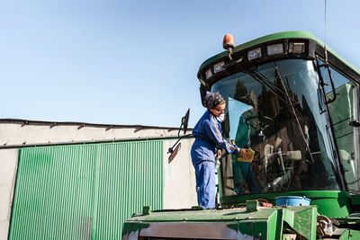 Side view low angle of busy female worker in uniform washing window of combine harvester parked in rural area