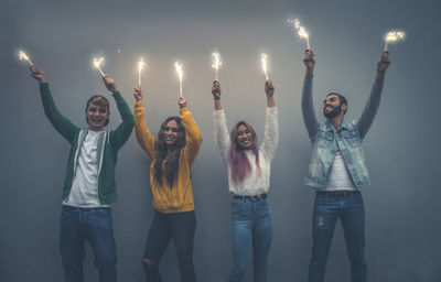 Portrait of cheerful friend holding sparkler sanding against wall