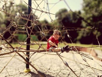 Full length of boy sitting on rope climbing frame at playground during sunny day