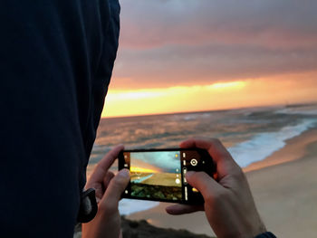 Midsection of man photographing sea against sky during sunset