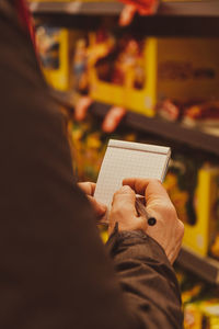 Female hands holding paper notebook of shopping list, blurred shelves on background. partial view of