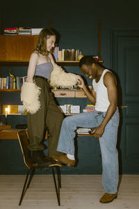 Happy young man holding hands of girlfriend standing on chair at home
