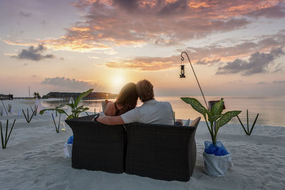 Rear view of couple sitting on sofa at beach during sunset