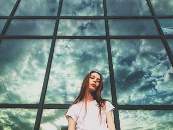 Low angle portrait of young woman against glass window with cloudy sky in background