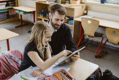 Smiling female student using tablet pc held by male teacher in classroom