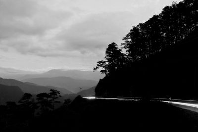 Silhouette trees by road against sky