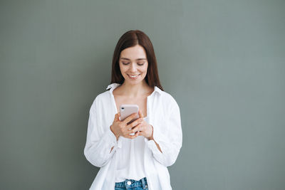 Portrait of smiling woman with dark long hair in white shirt using mobile phone on grey background