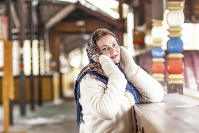 A slavic woman in a national colored scarf, a fur coat and  mittens on the porch of a wooden house