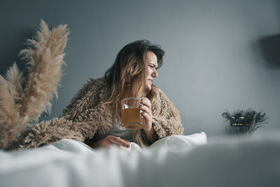 Young woman with herbal tea sitting on bed against wall