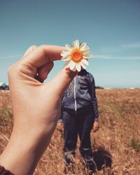 Cropped hand of woman holding flower against friend on field