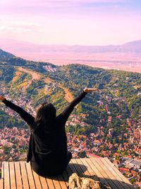 Rear view of young woman with arms raised looking at town while sitting on observation point