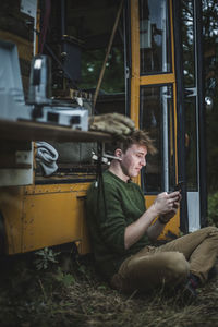 Full length side view of man sitting cross-legged while using smart phone against motor home during camping