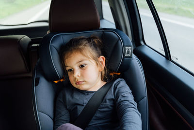 Thoughtful adorable tired little girl resting while sitting in car seat with fastened belt in modern vehicle during road trip