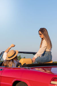 Two young women enjoying time together on a red classic convertible car on summer vacation time