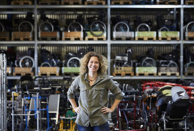 Portrait of smiling mature female worker standing with hands on hip against rack at warehouse