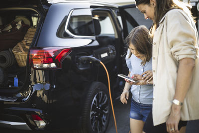 Portrait of mother, father and two daughters standing by car at electric vehicle charging station