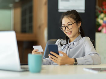 Woman holding visiting card while sitting by laptop on table in office