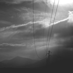 Electricity pylons on field with mountains against cloudy sky