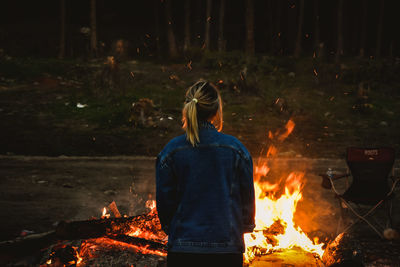 Rear view of woman standing by campfire in forest at night