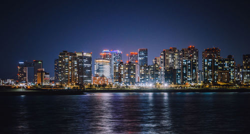 Illuminated buildings by river against sky at night