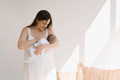 Adult woman embracing and pressing sleeping infant to chest gently against white wall