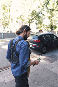 Mid adult father using mobile phone while carrying baby in carrier on sidewalk