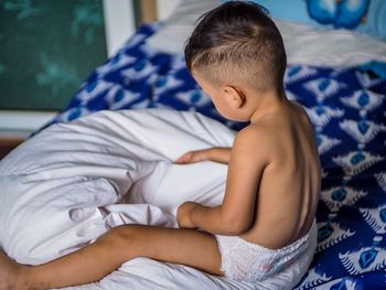 Side view of shirtless baby boy sitting on bed at home